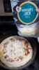 The breakfast pizza in the cast-iron pan. A bag of pre-shredded monterey jack cheese is being held above it, and a layer of the cheese covers the toppings.